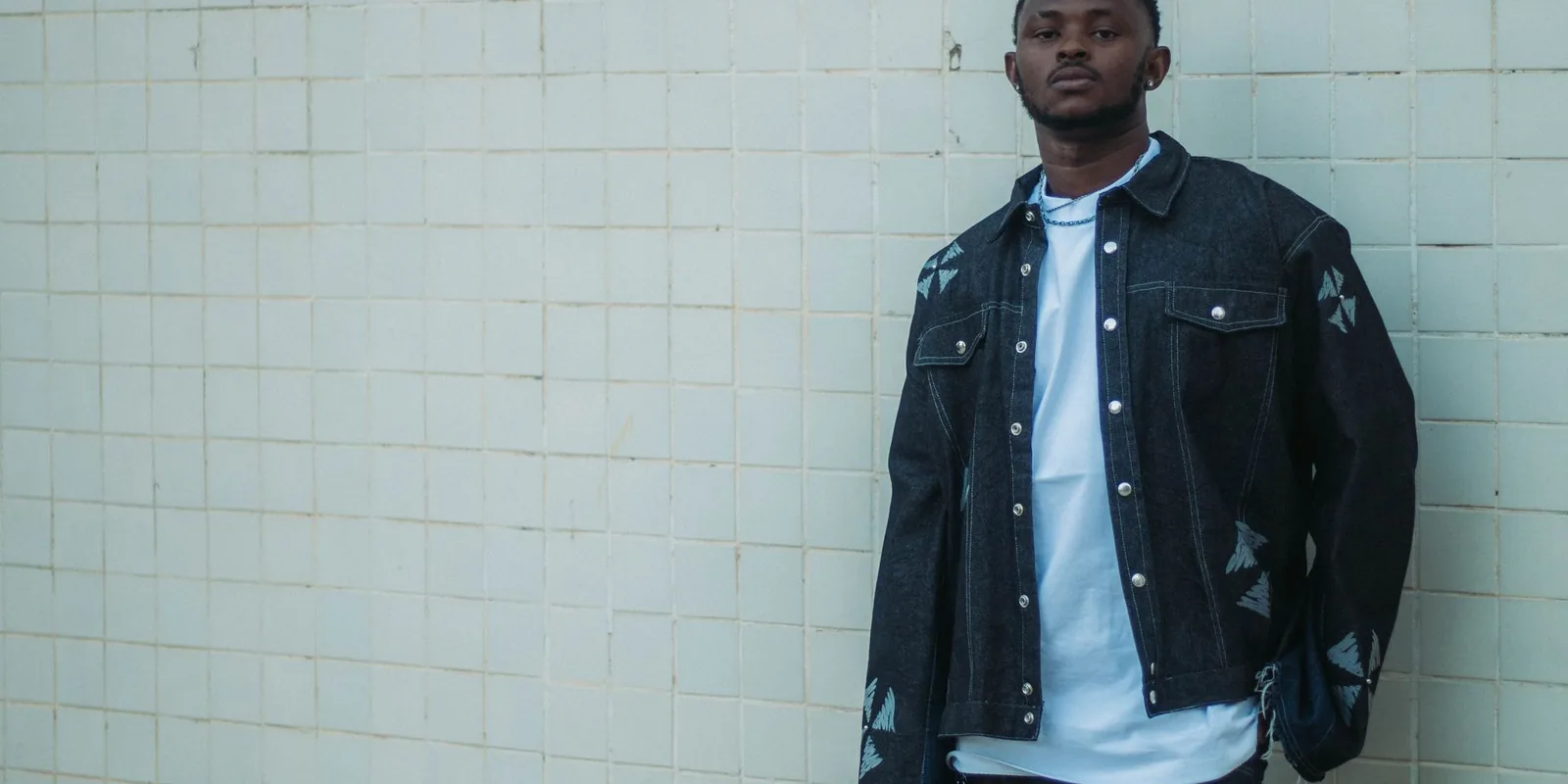 Fashionable man in a dark denim jacket and jeans poses against a tiled wall
