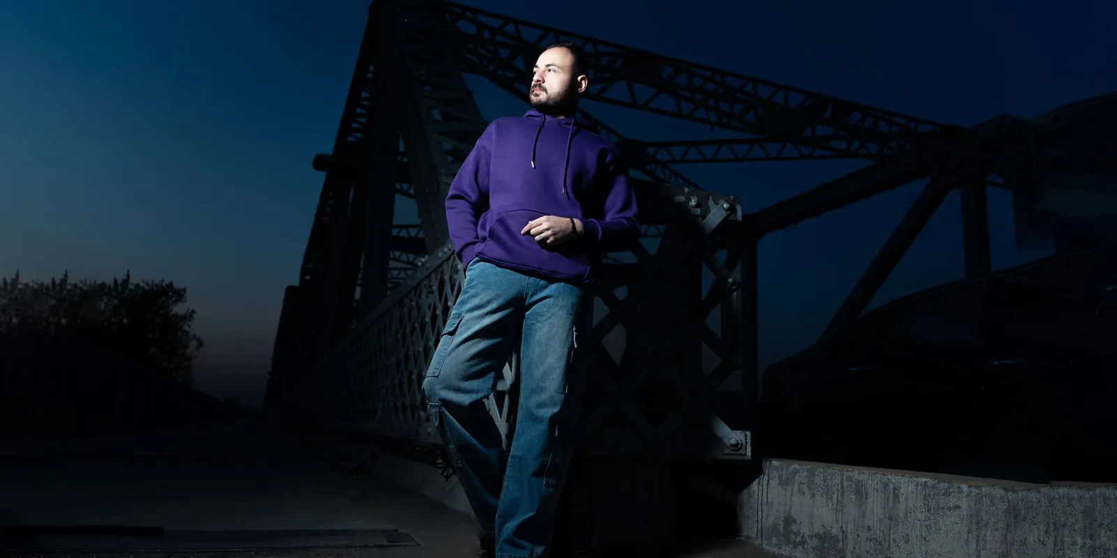 Man in casual attire poses confidently on a steel bridge at dusk