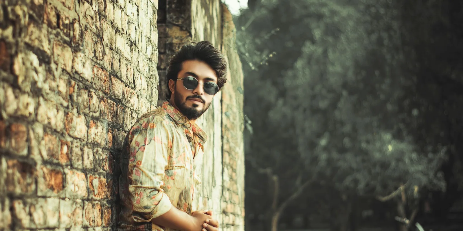 Man with sunglasses leaning against a brick wall, outdoors amidst trees