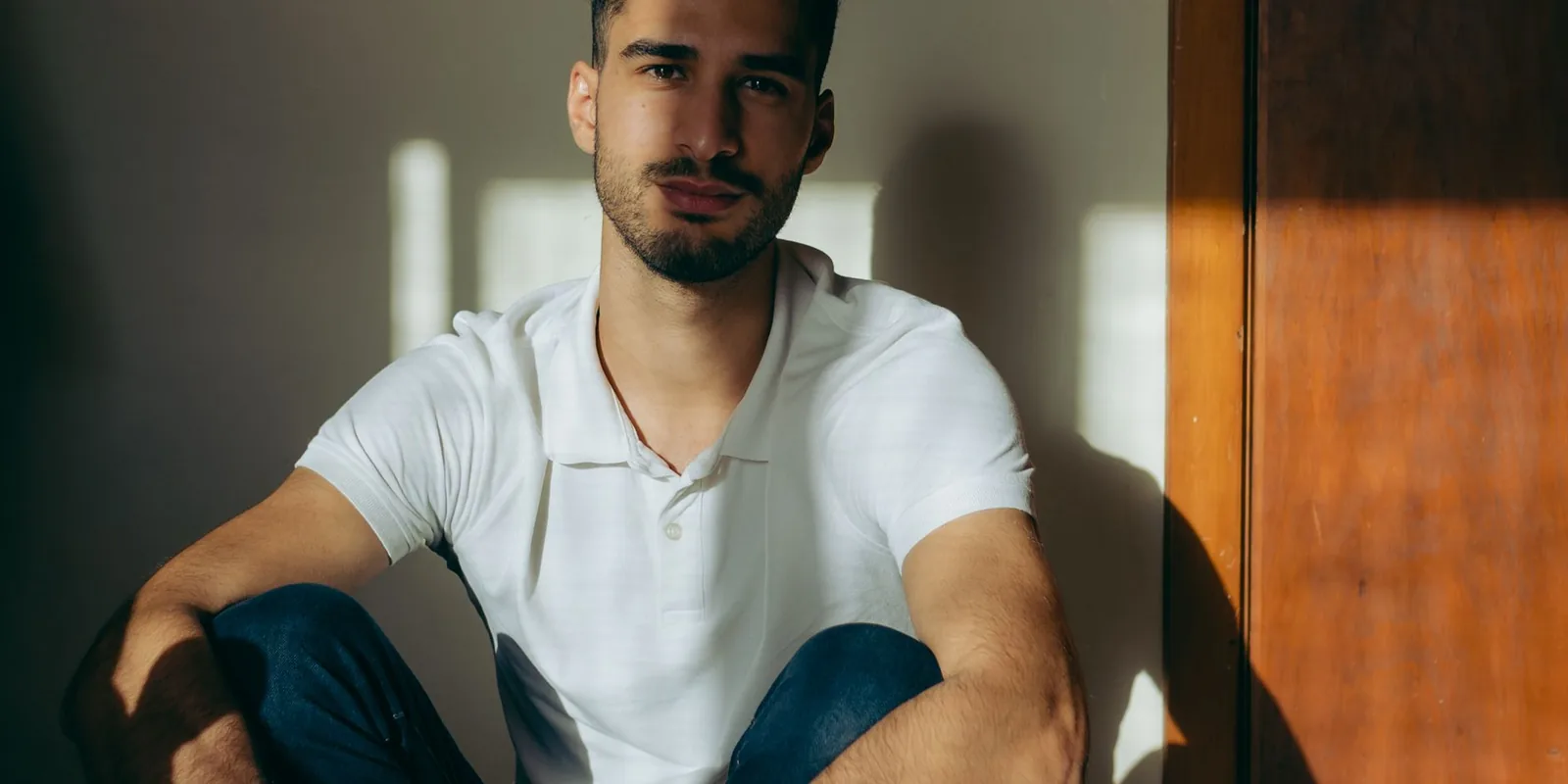 A young man in a white polo shirt sits indoors, looking at the camera with a serene expression