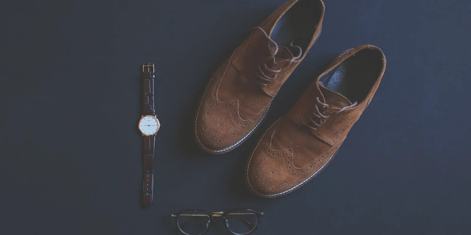 Flat lay of brown leather shoes, eyeglasses, and wristwatch on a dark background