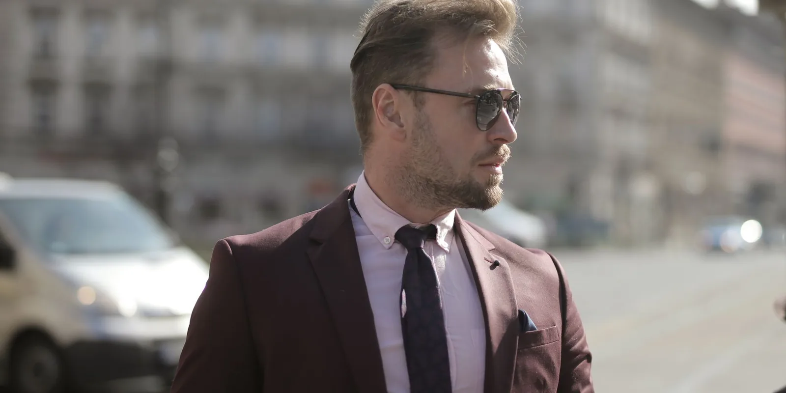 Businessman in sunglasses wearing maroon suit and tie walks confidently in urban setting