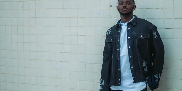 Fashionable man in a dark denim jacket and jeans poses against a tiled wall