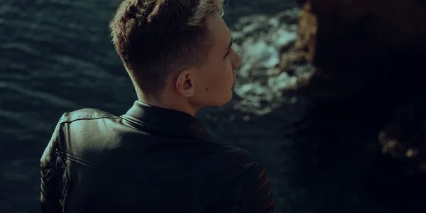 Young man in leather jacket looking thoughtfully over the sea from a rocky edge