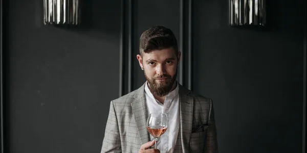 Bearded man in a gray blazer enjoying a glass of wine indoors. Elegant and sophisticated ambiance