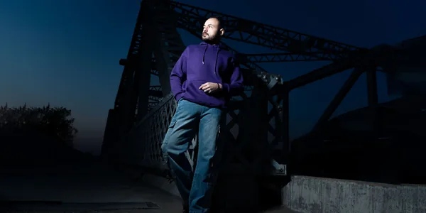 Man in casual attire poses confidently on a steel bridge at dusk