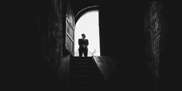 Black and white image of a man silhouetted against a bright doorway, standing on stairs