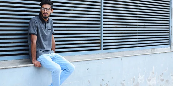 A young man in casual attire sitting by an urban wall with horizontal lines