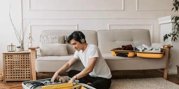 Young man in a living room, packing a suitcase while wearing headphones and listening to music