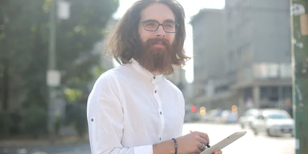 Bearded man wearing glasses and holding a tablet on a city street. Bright daytime setting