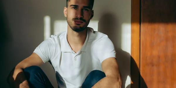 A young man in a white polo shirt sits indoors, looking at the camera with a serene expression
