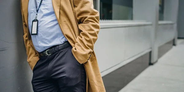 Crop anonymous male with hands in pocket in official outfit leaning on wall of building