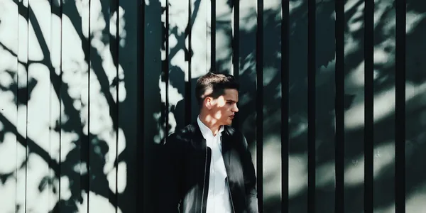 A young man in a black bomber jacket stands outdoors, with dramatic shadows